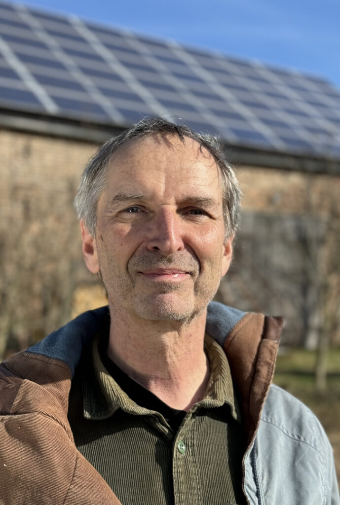 Ein Mann steht vor einem Dach auf dem eine Photovoltaik-Anlage verschwommen zu sehen ist und lächelt in die Kamera
