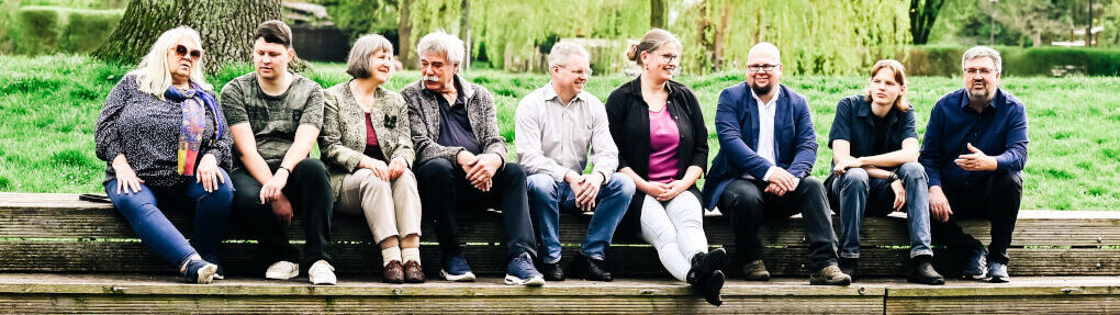 9 Personen sitzen nebeneinander auf einem Holzsteg und sprechen gut gelaunt miteinander